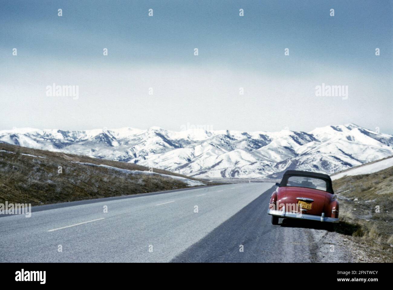 Long straight road with 1952 red Plymouth convertible car parked at the roadside with a view of snow covered Rocky Mountains range, USA in the 1950s Stock Photo