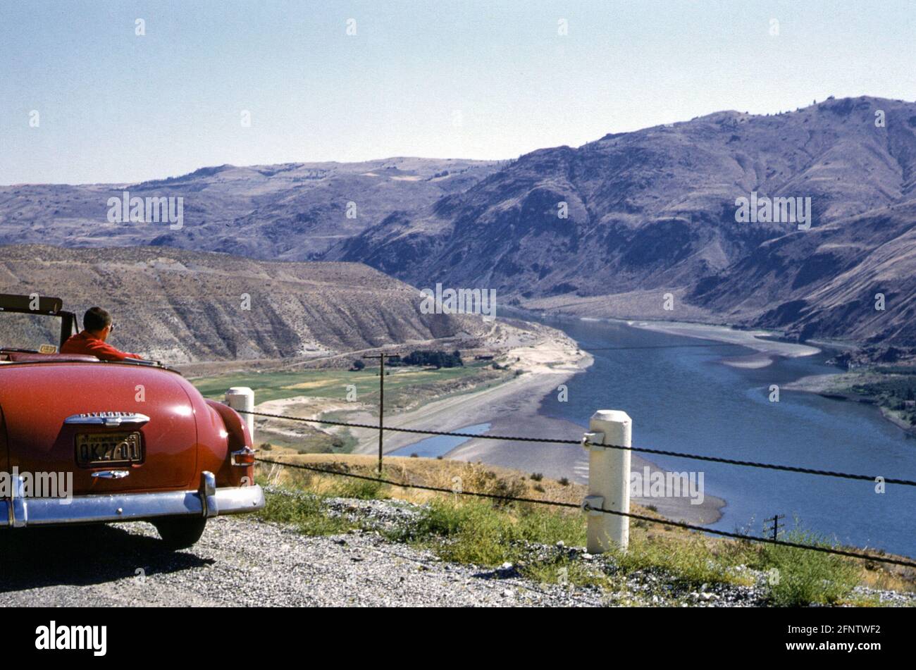 Young man sitting in a red 1952 Plymouth convertible car with the top down in the 1950s. The car is parked at the roadside overlooking the Columbia River, Washington state, USA on a sunny day Stock Photo