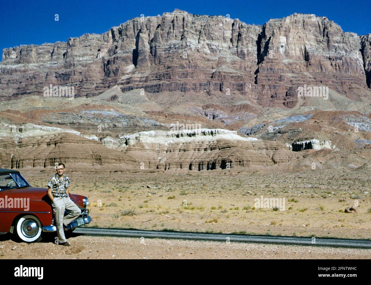 Young man leaning on a red 1952 Plymouth convertible car with white wheel rims in 1950s. The car is parked at the roadside next to the Painted Desert cliffs, Arizona, USA in the 1950s Stock Photo
