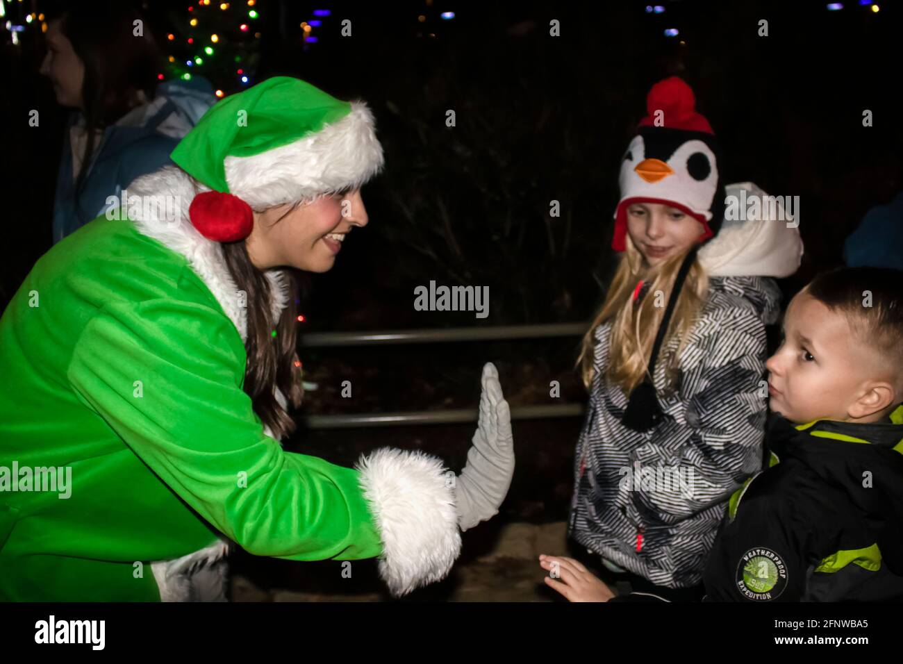 12-15 2018 Tulsa USA Female employee dressed as green elf high fives unsure little boy at Gathering Place public park at nighttime festivities -select Stock Photo