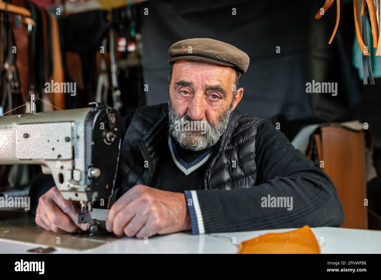 Old bearded man with serious face looking at the camera while using sewing machine working with leather. Stock Photo