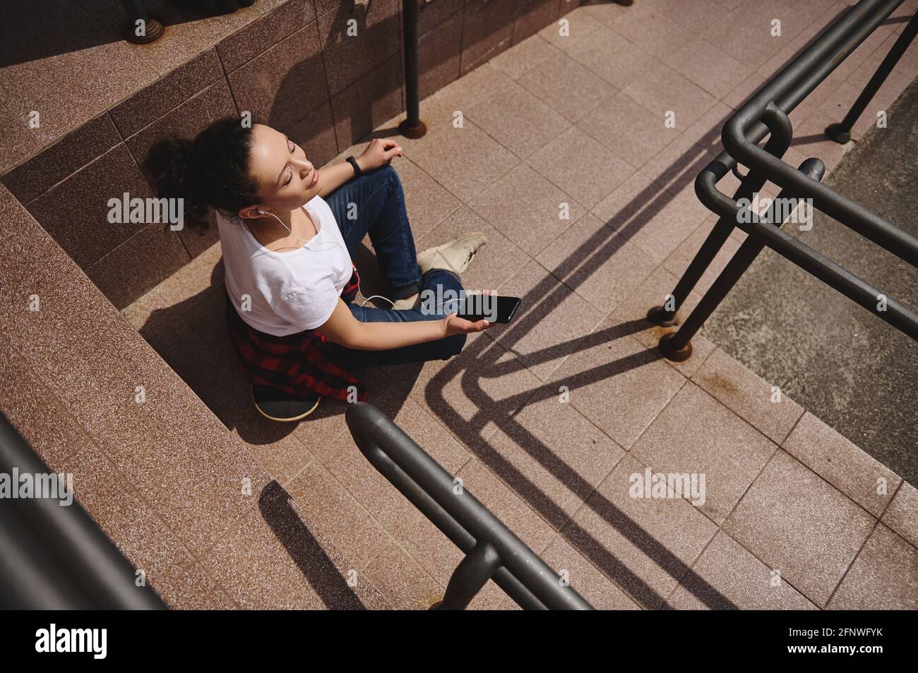 High angle view of a woman relaxing sitting on skateboard with headphones and smartphone in her hand and taking sun bathing outdoors Stock Photo