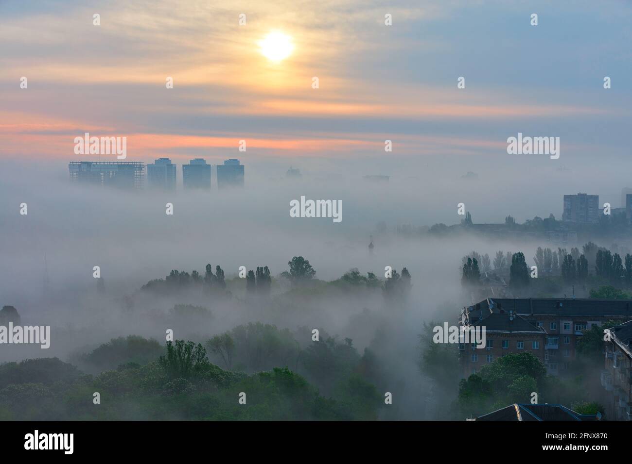 A thick fog, like a duvet, spreads early in the morning between the houses and trees of the city park in the soft touch of the rising sun. Stock Photo