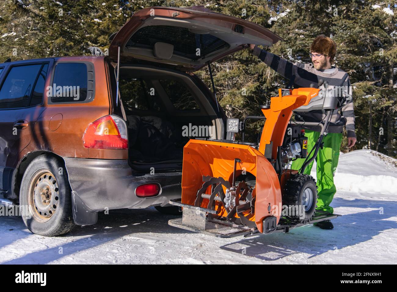 Wide angle shot of a bright orange mechanical snowplough being downloaded from the trunk of a car and ready to be used for fresh snow removal. Stock Photo