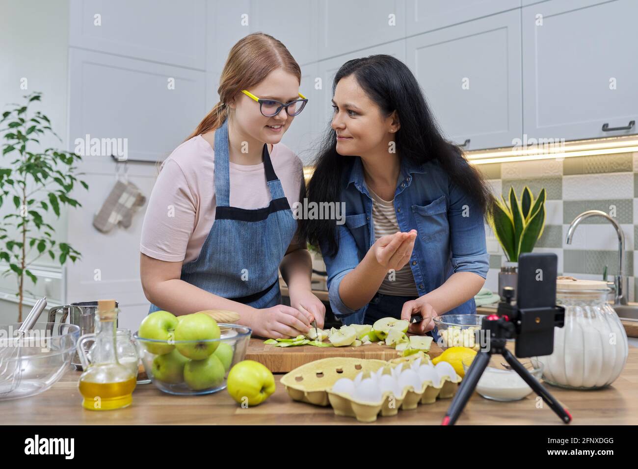 Mom and teen daughter cooking apple pie together, looking at smartphone screen Stock Photo