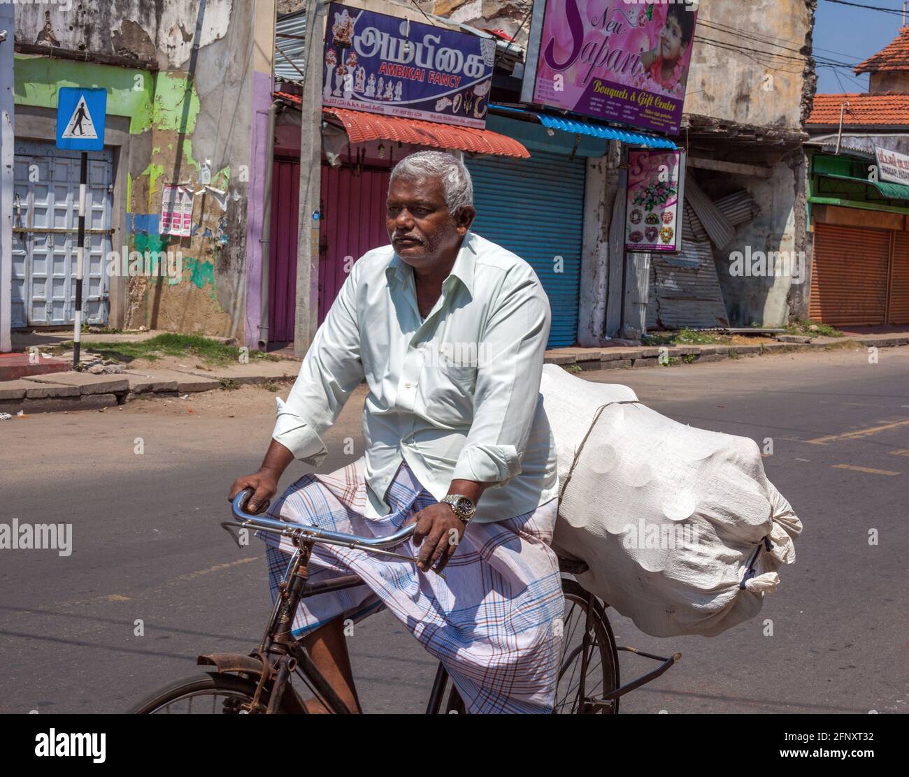 Elderly Sri Lankan male wearing lunghi rides bicycle overloaded with package on rear, Jaffna, Northern Province, Sri Lanka Stock Photo