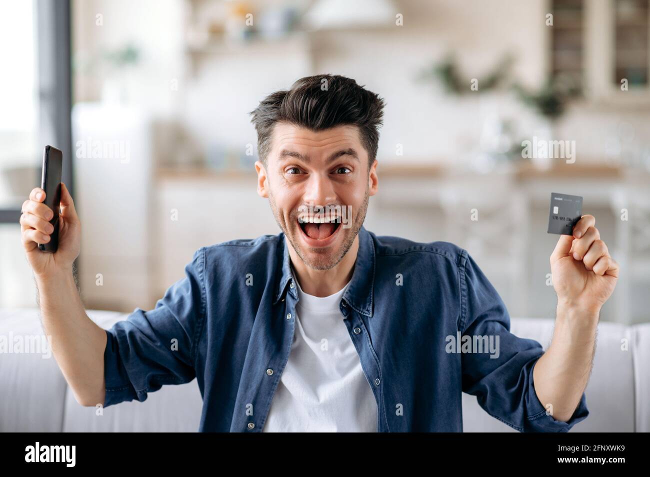 Amazed positive caucasian unshaven guy in a denim shirt, holds his smartphone in one hand and a credit card in the other, made an online payment transaction, shopping online, happy with good shopping Stock Photo