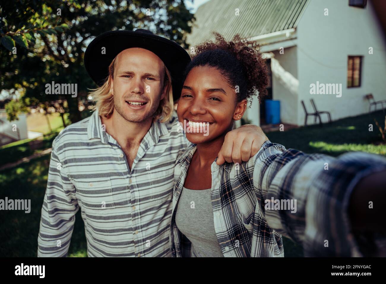 Mixed race couple smiling taking selfie with cellular device standing in front of white farm home Stock Photo