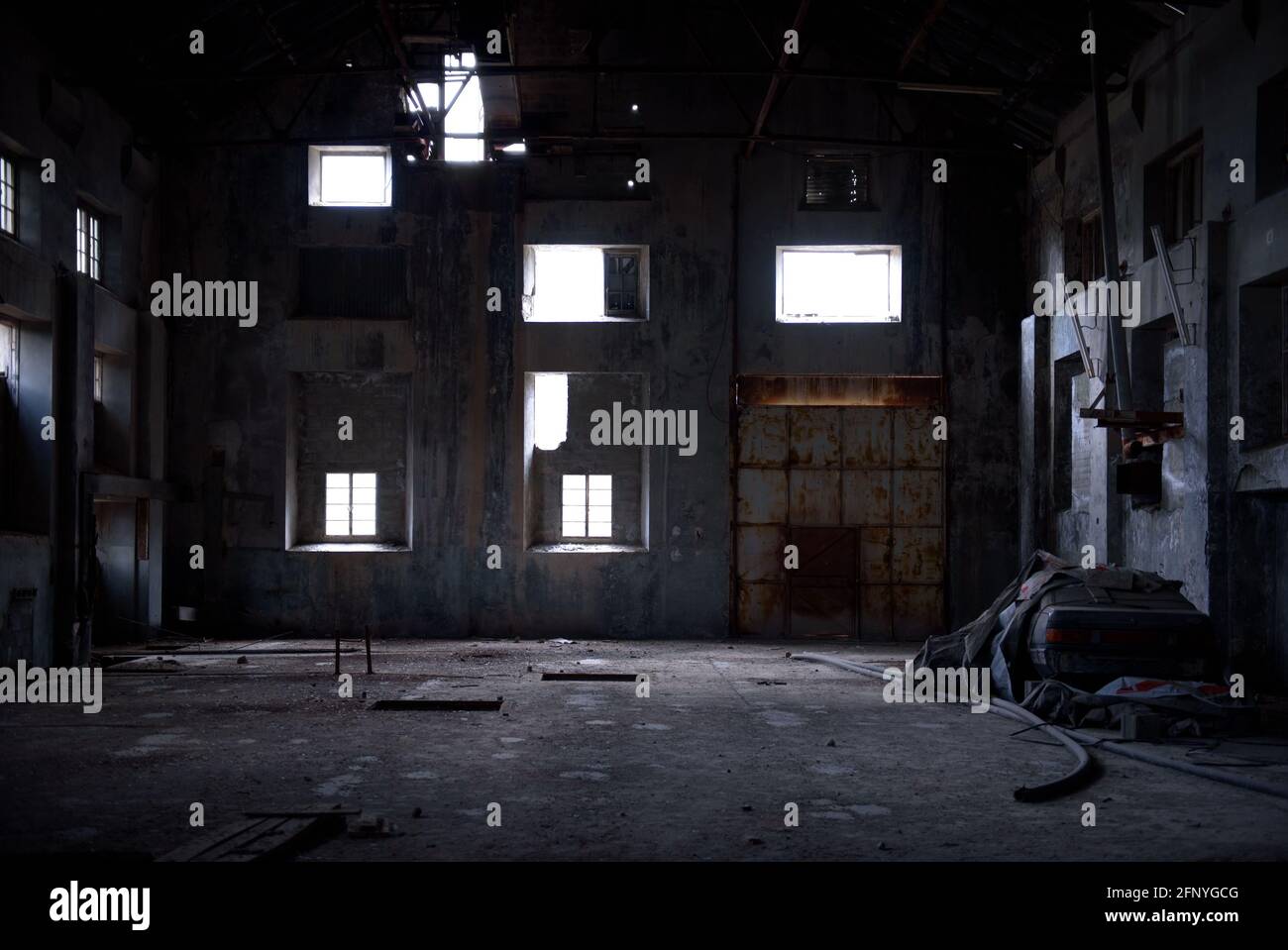 Abandoned industrial backdrop. Old dark warehouse interior with concrete walls, rusty gate and light falling through broken windows Stock Photo