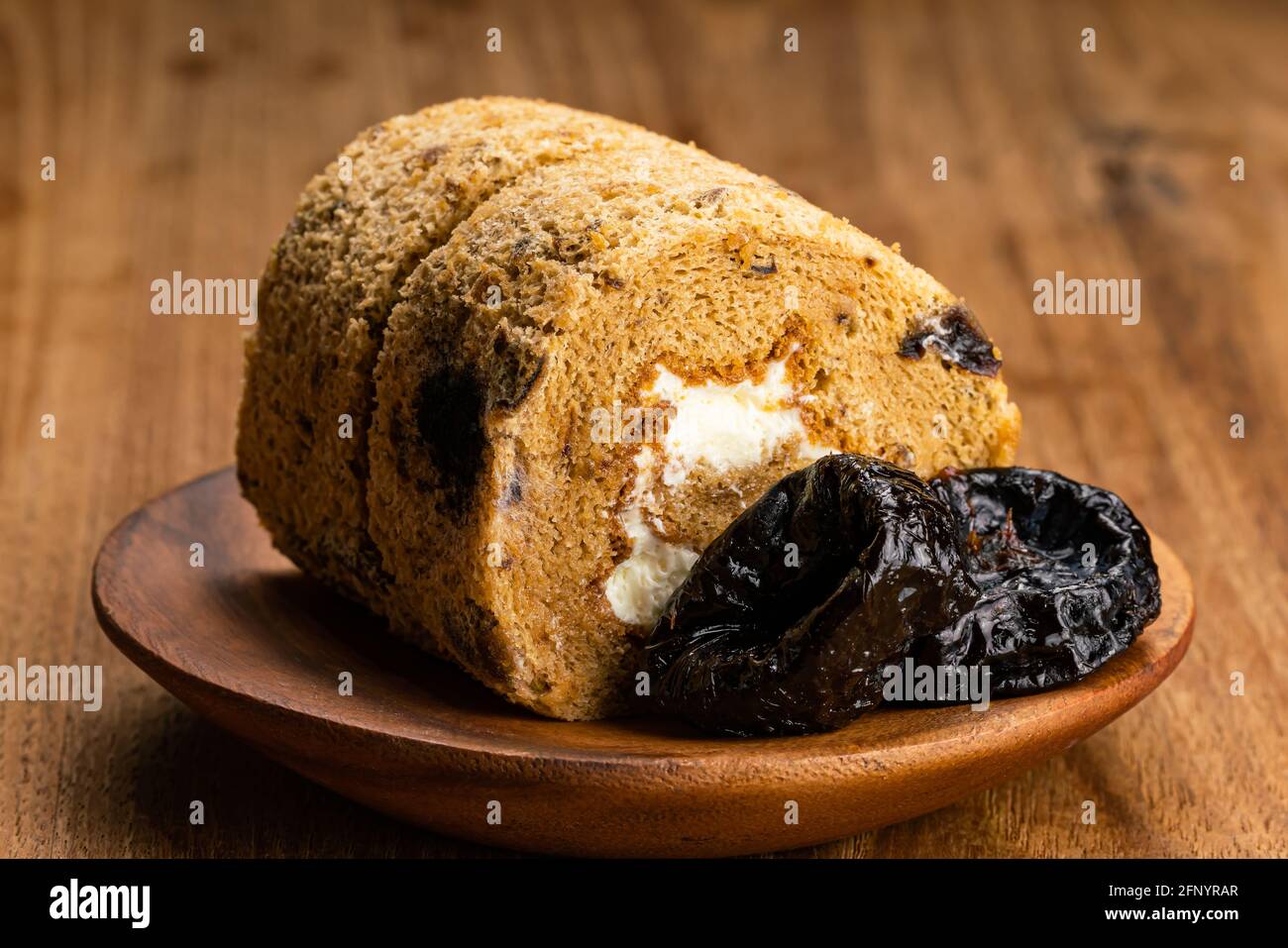 Closeup view of homemade prune sponge cake roll and dried pitted prune fruits in wooden plate on wooden table. Stock Photo