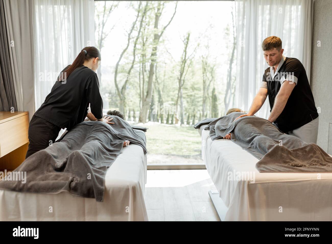Two masseurs doing a deep back massage to guests with pampering technique. Wellness and leisure time for health care together with forest view in window. Stock Photo