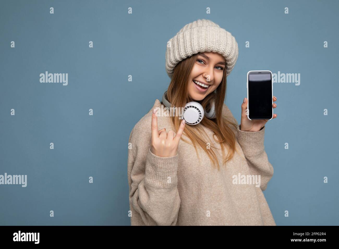 Photo of beautiful positive joyful young dark blonde woman wearing beige stylish sweater and knitted beige winter hat isolated on blue background Stock Photo