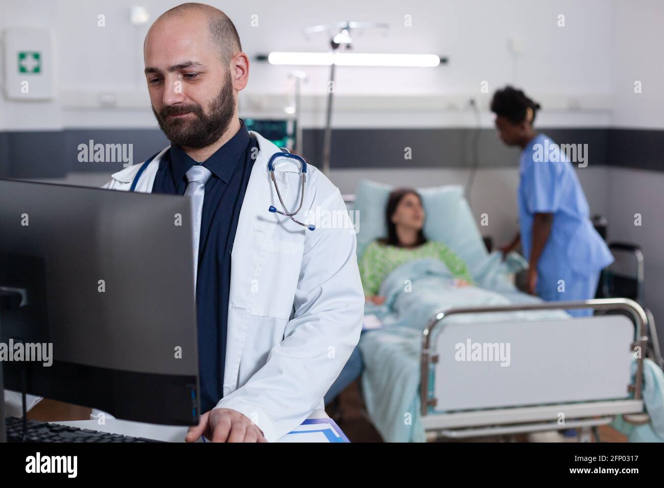 Front view of practitioner doctor typing medical expertise on computer while in background black asisstant discussing healthcare treatment. Hospitalized patient having respiratory disorder Stock Photo