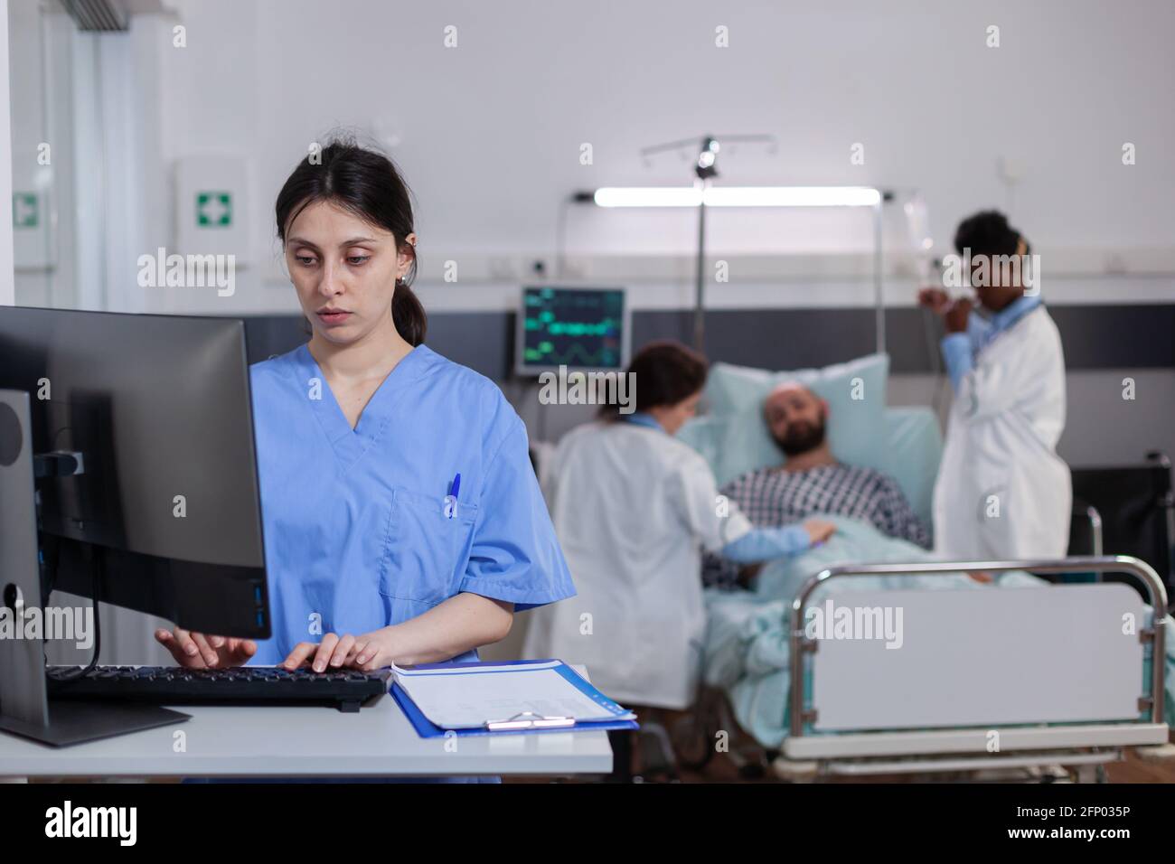 Front view of medical asisstant typing illness expertise on computer while in background physician doctors discussing with sick man. Hospitalized patient having respiratory disorder Stock Photo