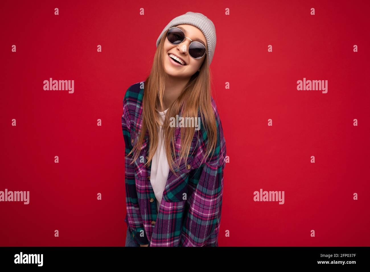 Photo shot of beautiful smiling young dark blonde woman isolated over red background wearing purple shirt grey hat and stylish sunglasses looking at Stock Photo