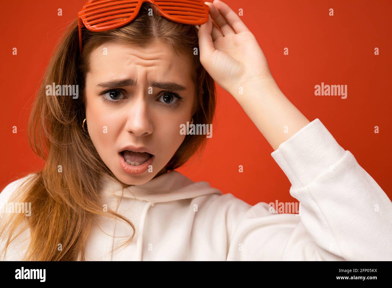 Closeup photo shot of beautiful sad shocked young blonde woman isolated over colourful background wearing casual white hoodie and stylish glasses Stock Photo