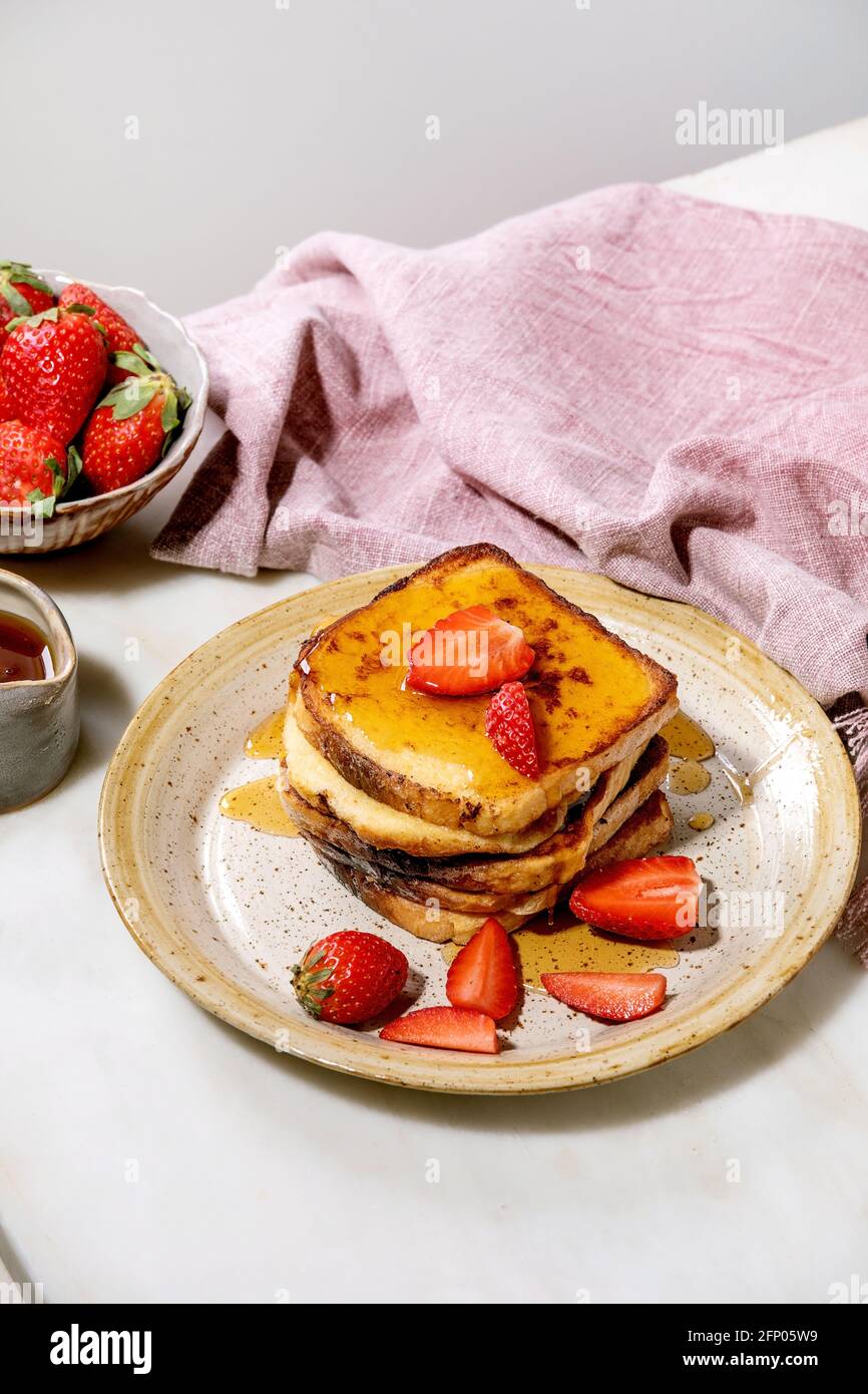 Stockpile of french toasts with fresh strawberries on ceramic plate, maple syrup in ceramic jug and pink cloth napkin over white table. Home breakfast Stock Photo