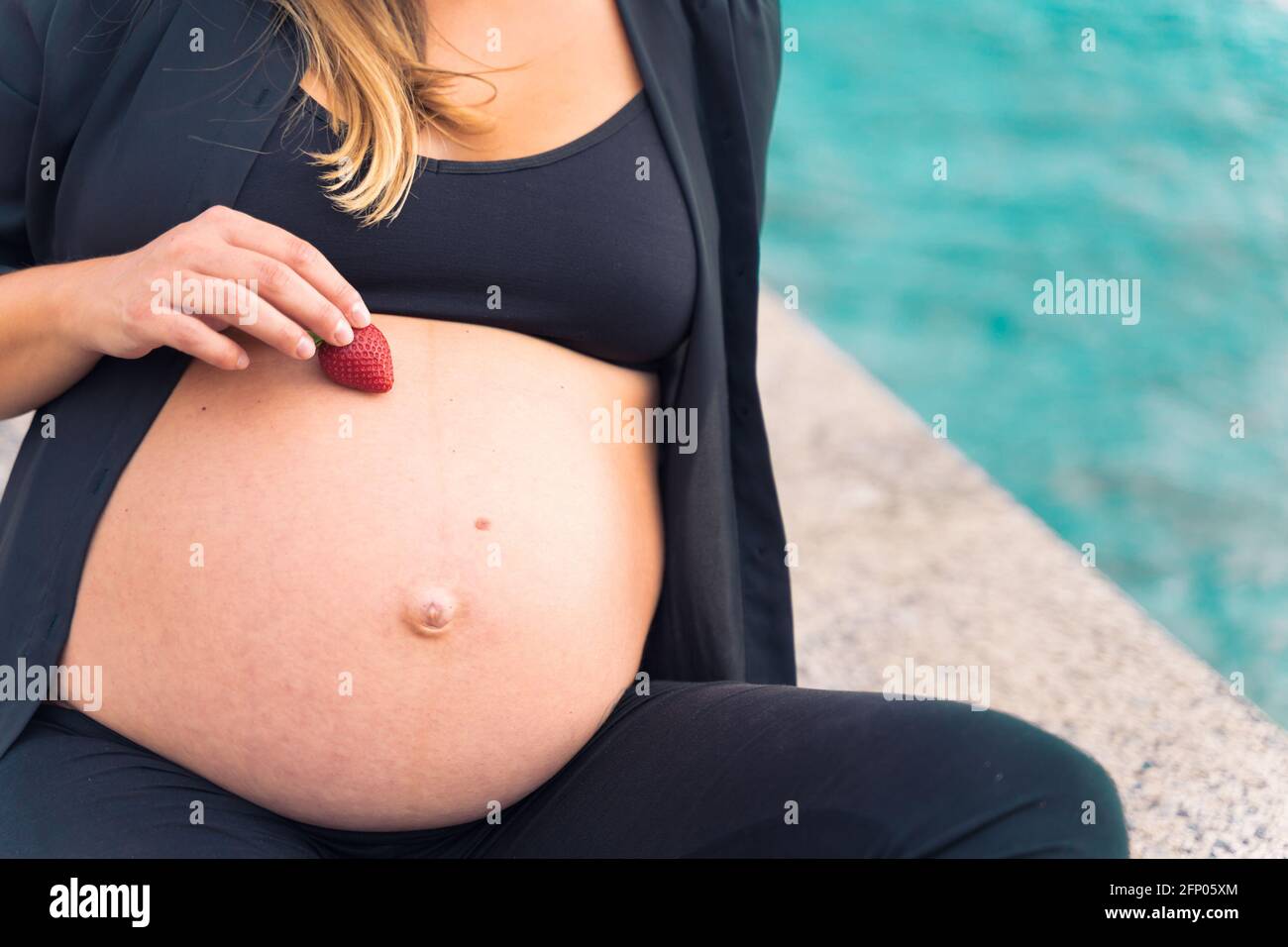 A pregnant in sportswear woman holding fresh delicious strawberry near of sea. Copy space. Berrys, alergia, diet and healthy food concept. Stock Photo