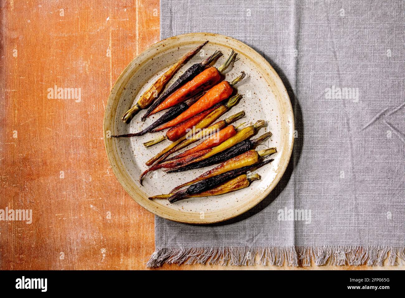 Grilled different colored carrots on ceramic plate with grey textile napkin over orange stone background. Top view, flat lay. Copy space. Vegan dinner Stock Photo