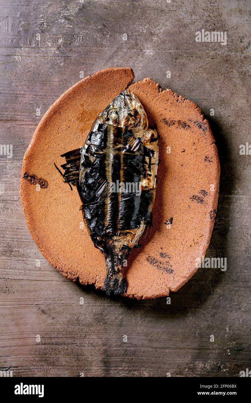 Grilled cooked fresh gutted sea bream or dorado fish on ceramic plate wrapped in bamboo leaves over dark brown texture background. Top view, flat lay Stock Photo