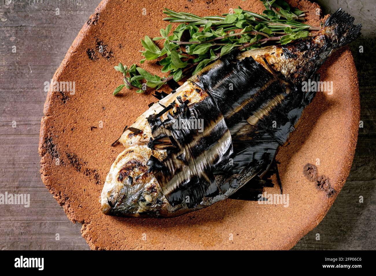 Grilled cooked fresh gutted sea bream or dorado fish on ceramic plate wrapped in bamboo leaves served with herbs over dark brown texture background. T Stock Photo