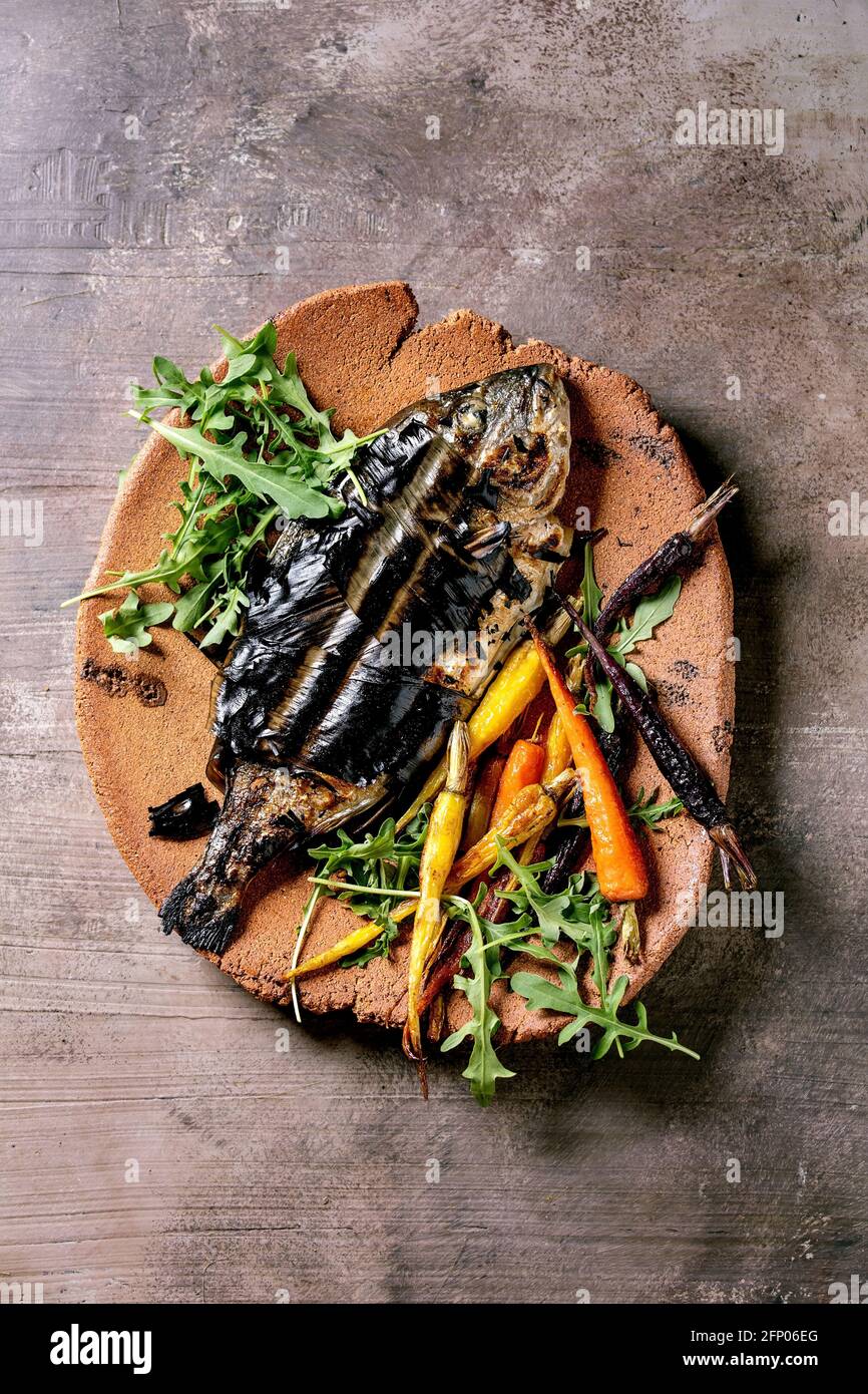 Grilled cooked fresh gutted sea bream or dorado fish on ceramic plate wrapped in bamboo leaves served with herbs, colorful carrots over dark brown tex Stock Photo