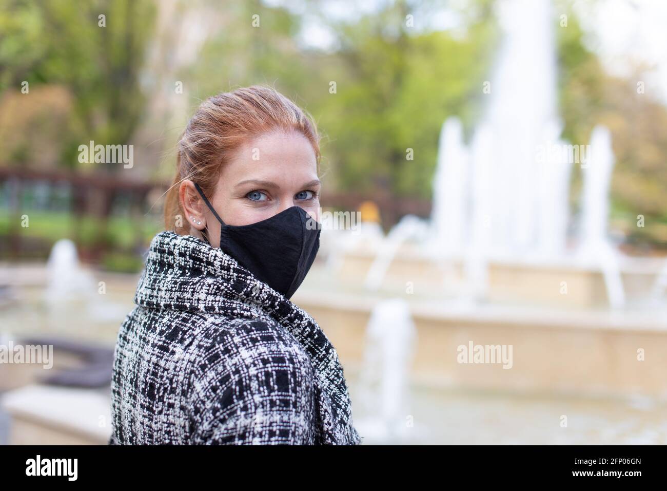 Young woman in black mask looking back at public park with fountain Stock Photo