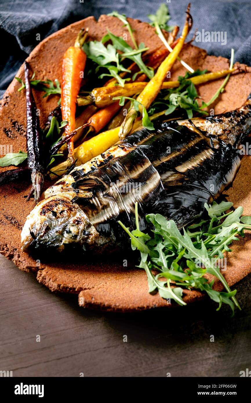 Grilled cooked fresh gutted sea bream or dorado fish on ceramic plate wrapped in bamboo leaves served with herbs, colorful carrots, blue napkin over d Stock Photo