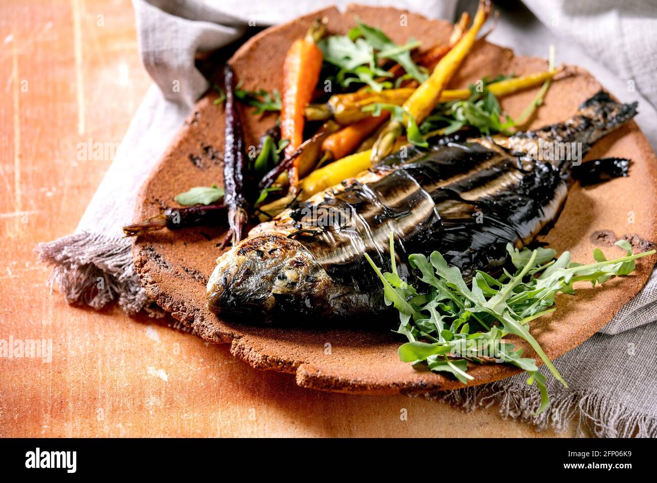 Grilled cooked fresh gutted sea bream or dorado fish on ceramic plate wrapped in bamboo leaves served with herbs, colorful carrots, white napkin over Stock Photo