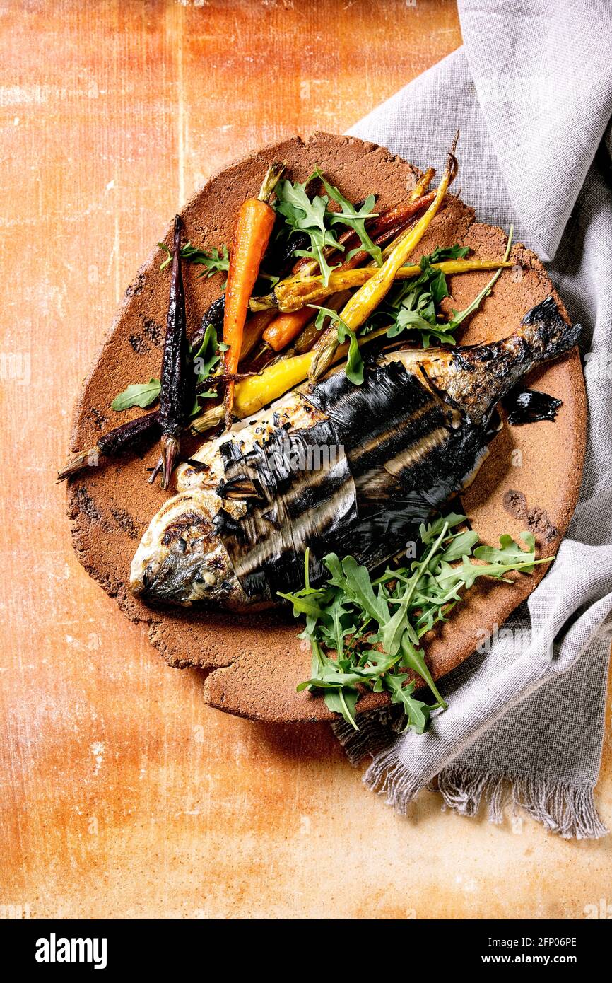 Grilled cooked fresh gutted sea bream or dorado fish on ceramic plate wrapped in bamboo leaves served with herbs, colorful carrots, white napkin over Stock Photo