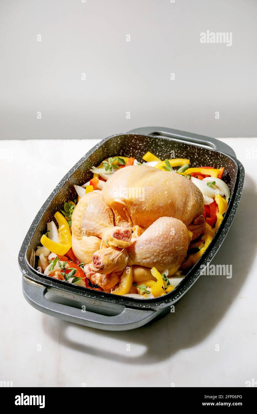 Raw organic uncooked whole chicken with sliced vegetables bell pepper, onion and herbs in oven tray, ready to cook. White marble table. Stock Photo