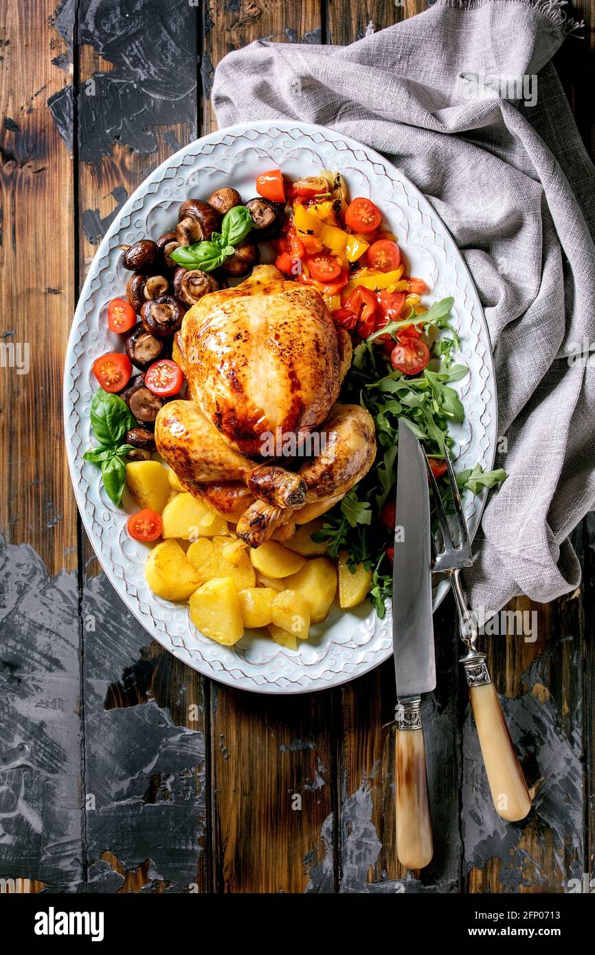 Grilled cooked whole chicken with vegetable garnish grilled bell pepper, onion, baked potatoes, cherry tomatoes, mushrooms and herbs in ceramic plate Stock Photo