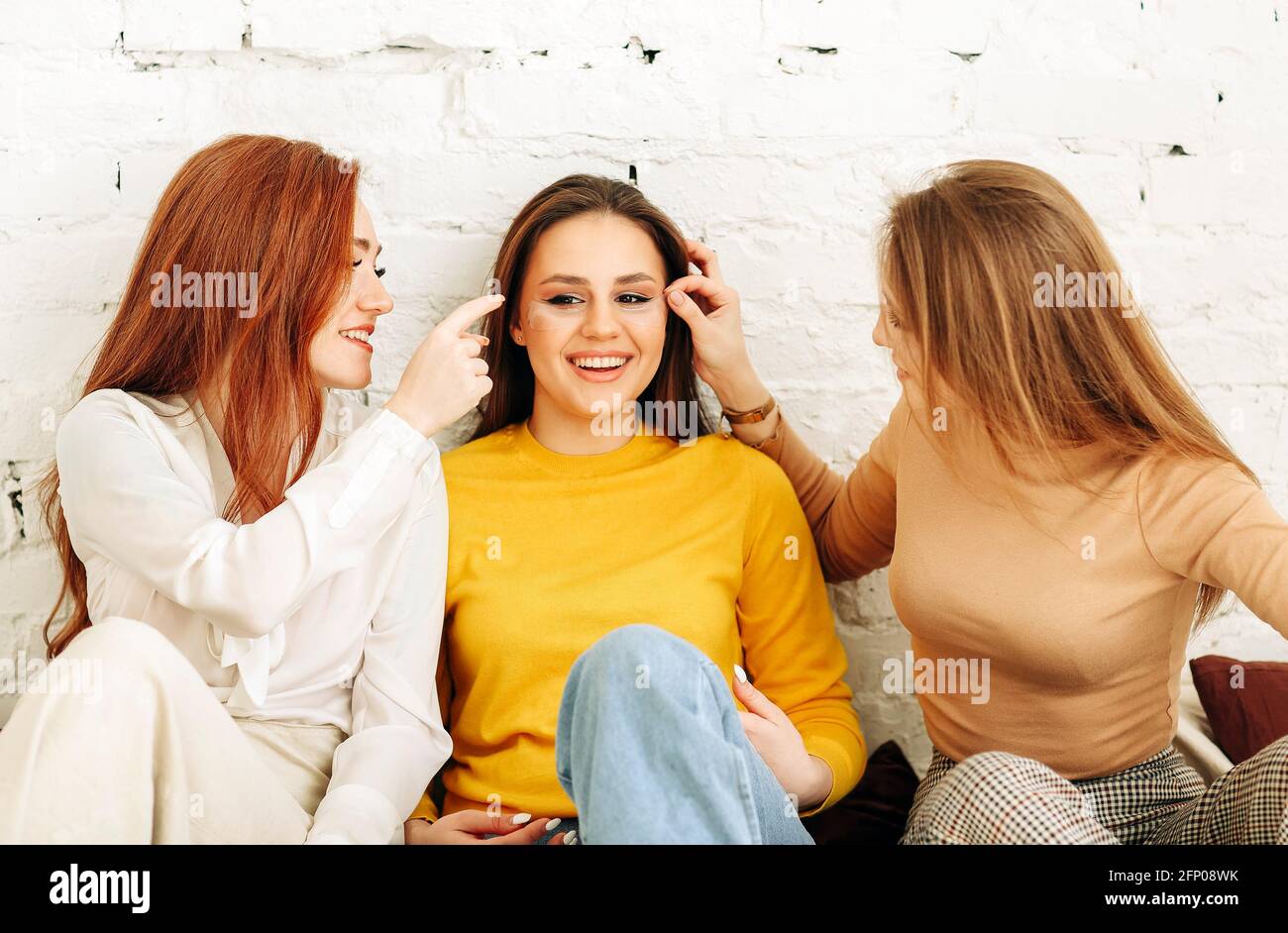 Optimistic young women smiling and touching face of stylish girlfriend while resting near rough brick wall Stock Photo