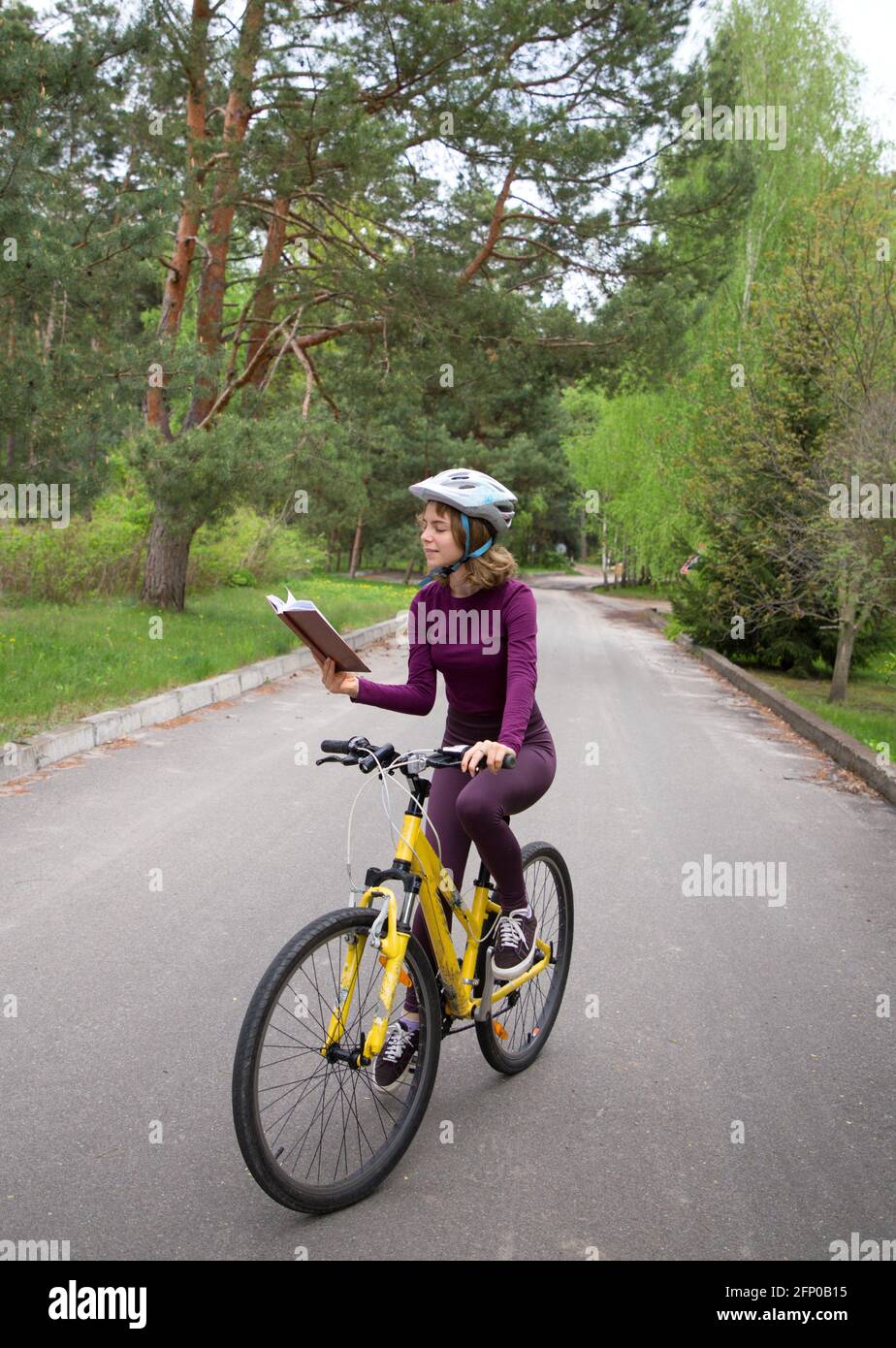 pretty girl teenager in a helmet rides a bicycle and reads a book with enthusiasm in motion. Combine the hobby of reading and cycling. Digital detox Stock Photo