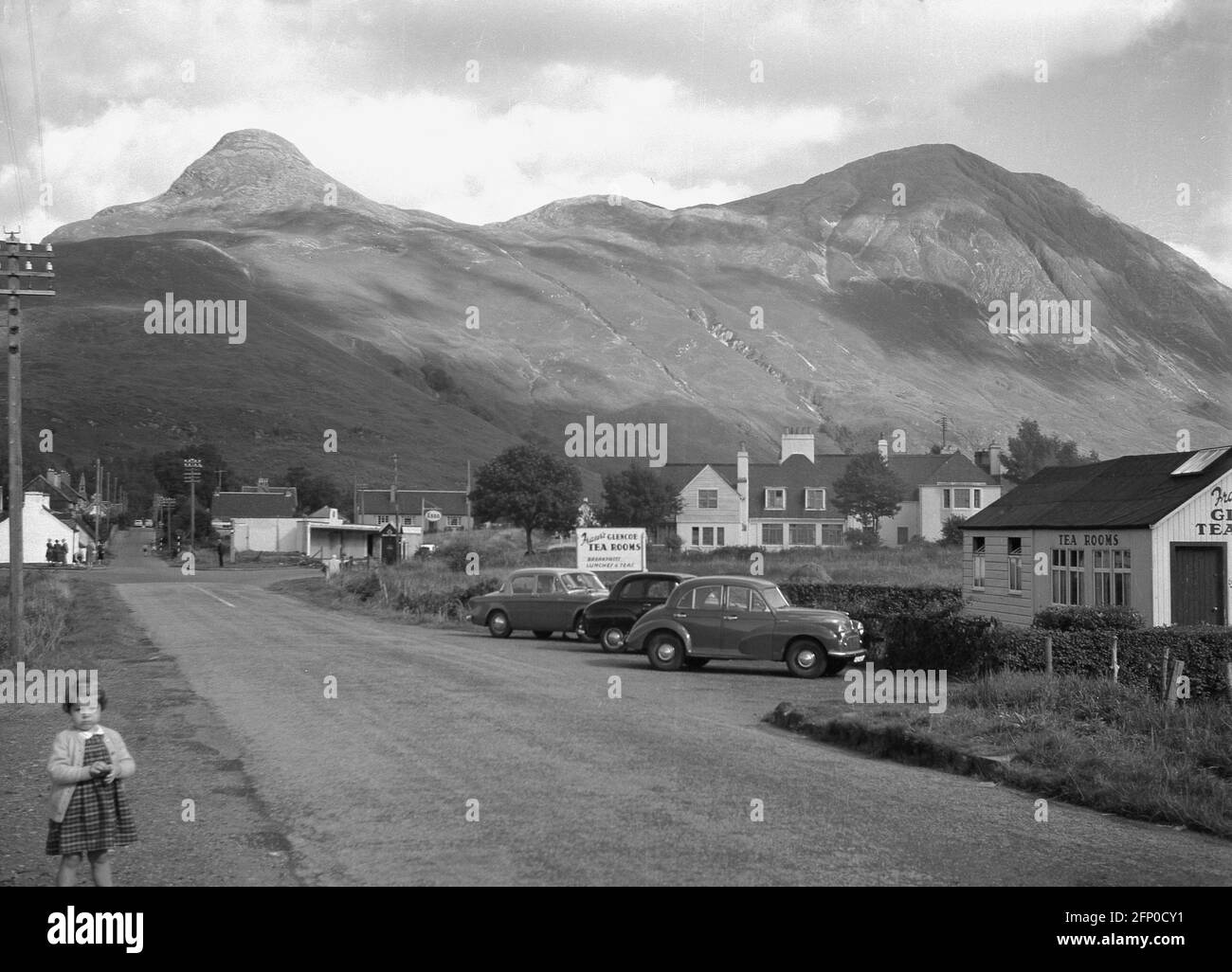 1960, historical, cars of the era parked outside Fraser's Tea rooms in the village of Glencoe, Highlands, Scotland, UK with the scottish mountains of Pap of Glenccoe in the background. Glen Coe is one of Scotland's most famous Highland glens and is known as the gateway to the Highlands. Stock Photo