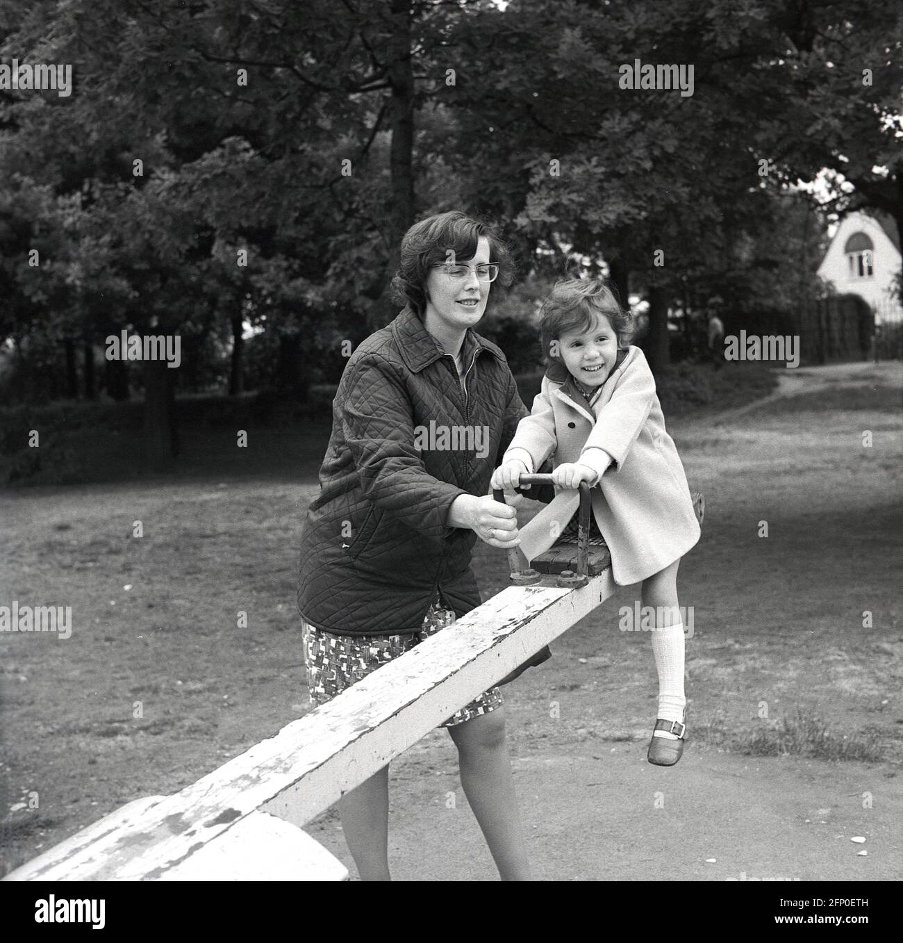 1960s, historical, with her mother beside her, a young girl on a metal see-saw at a park playground, England, UK. Also known as a teeter-totter or teeterboard. A traditional piece of playground equipment, the seesaw is a long narrow metal, as seen here (or wooden) beam with a pivot point in the centre,  so as one end goes up, the other goes down, with handles eiither end to hold onto. Great fun, they were a common site in public playgrounds across Britain in this era Stock Photo