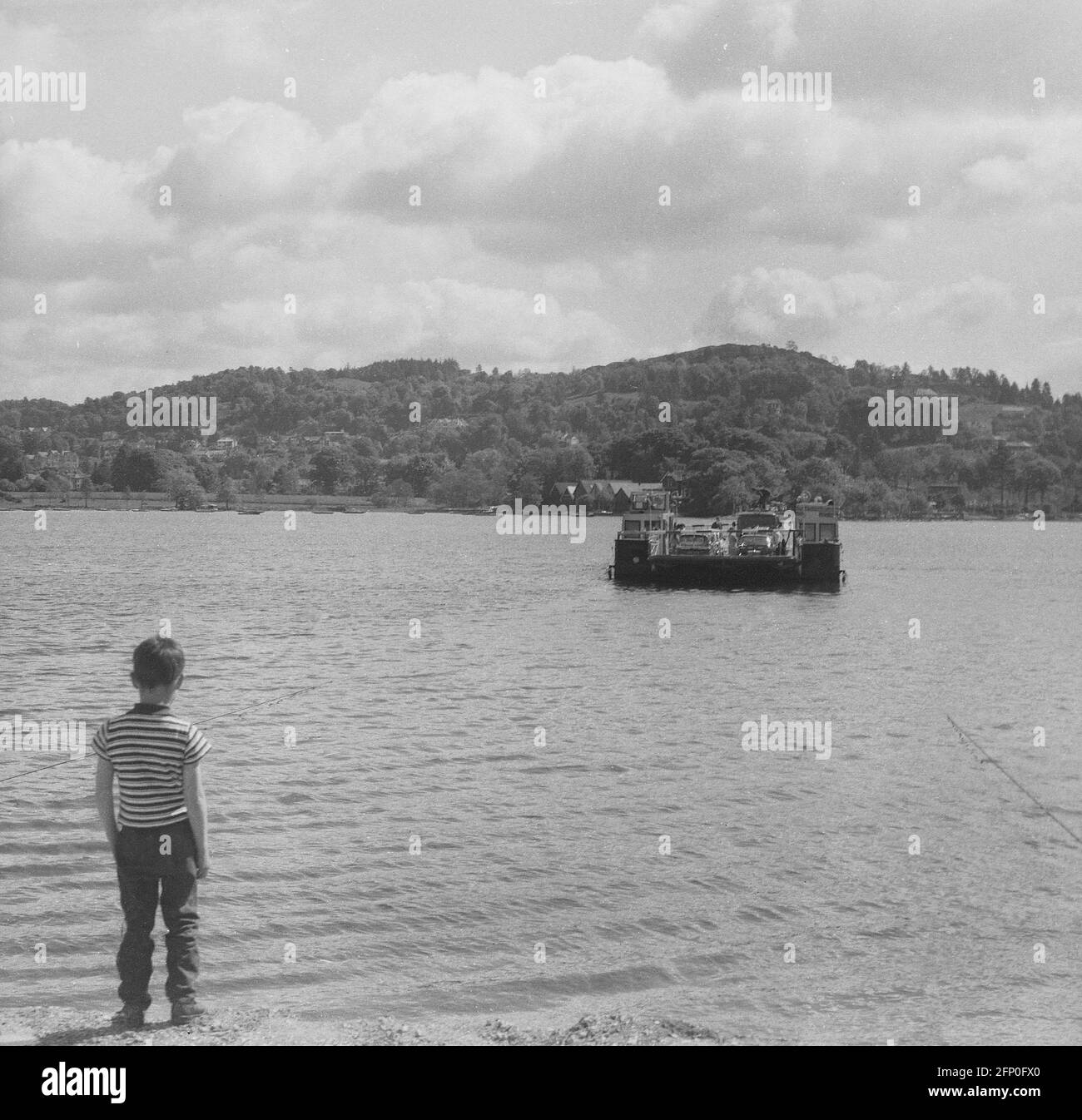 1961, historical, a young boy standing shoreside beside the calm water of a loch, watching a small cable or chain ferry taking cars and people going across the lake, Highlands of Scotland. There are hundreds of small off-shore islands in Scotland and some of the remote ones can only be reached by ferry. A cable ferry goes across a body of water pulled by cables connected to both shores, early cable ferries used rope or steel chains, hence the name of chain ferry. Stock Photo