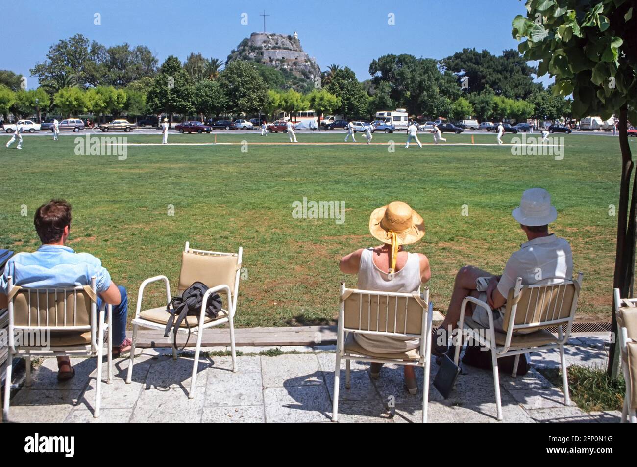 People watch cricket match in historical Liston cricket ground landscape part of Spianada Square Greek Corfu Town with Old Fortress beyond Greece EU Stock Photo