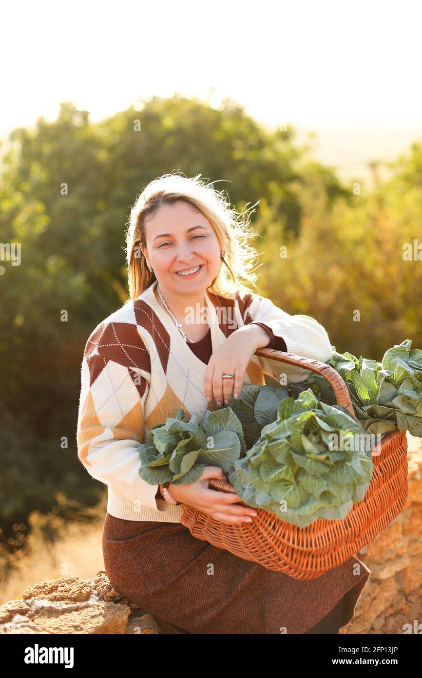 female leaning on wicker basket with fresh cabbage and touching face while resting on brick fence on sunny summer day in countryside Stock Photo