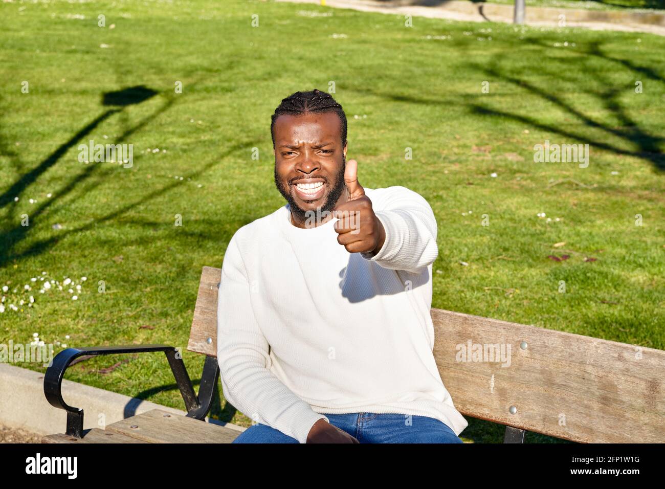 Casual African American young man sitting on park bench and showing thumbs up gesture to camera. High quality photo Stock Photo