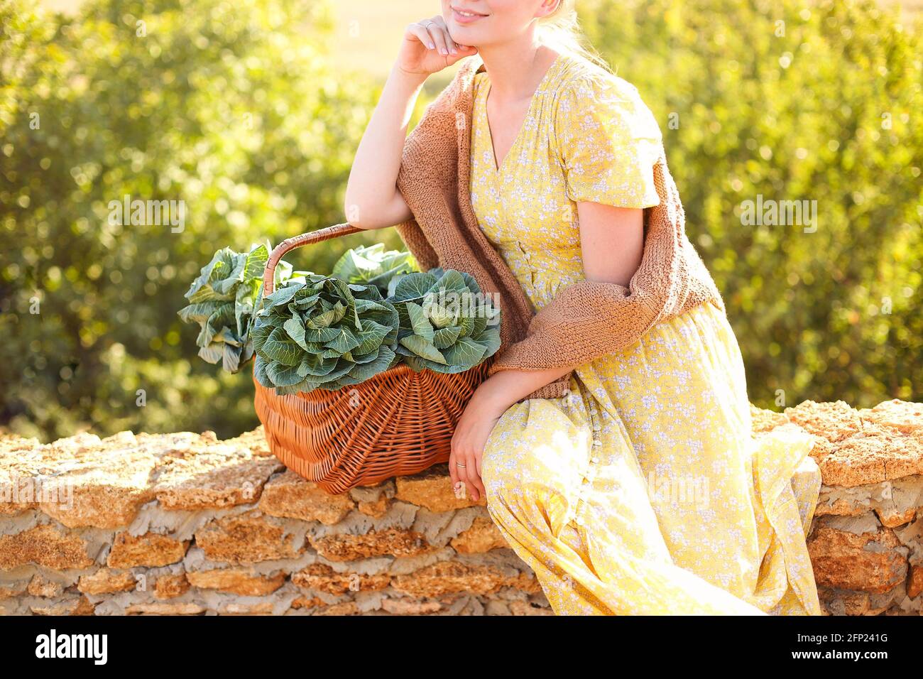 Unrecognizable young female leaning on wicker basket with fresh cabbage and touching face while resting on brick fence on sunny summer day in countrys Stock Photo
