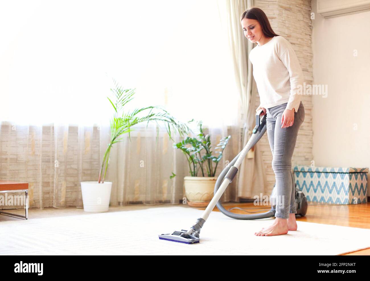 Full body young female using vacuum cleaner to tidy carpet in light room at home Stock Photo