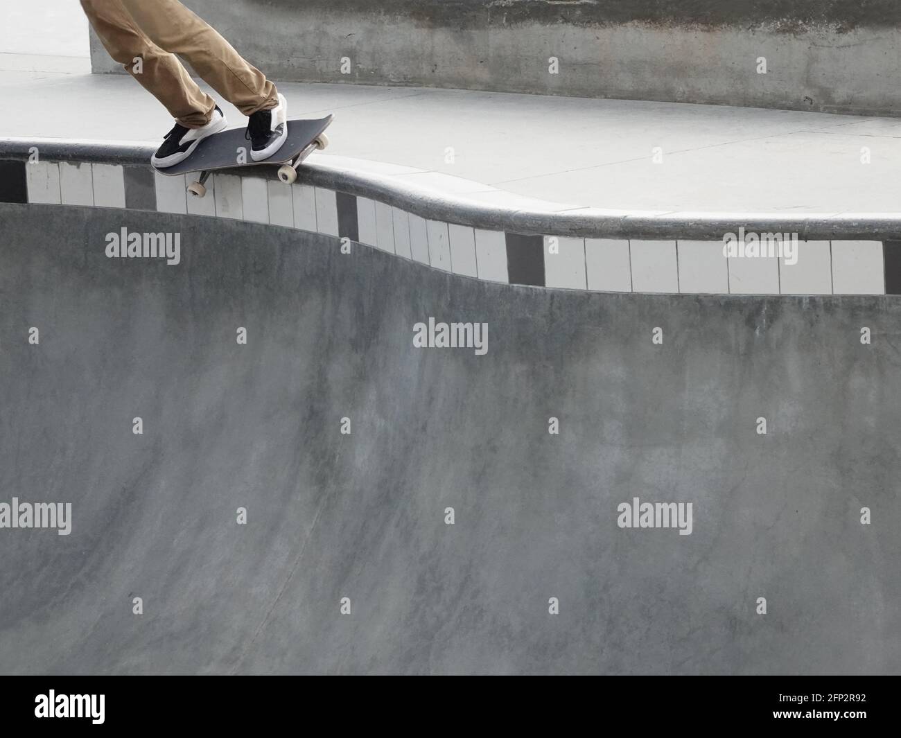 The legs and feet of a skateboarder are shown as the skateboard trucks grind the rail along a edge of a steep concrete bowl at a skate park. Stock Photo