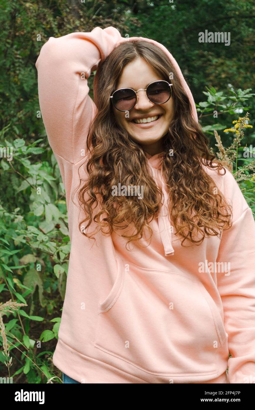 A young woman in a pink hoodie in sunglasses, with curly hair, smiles. Stock Photo