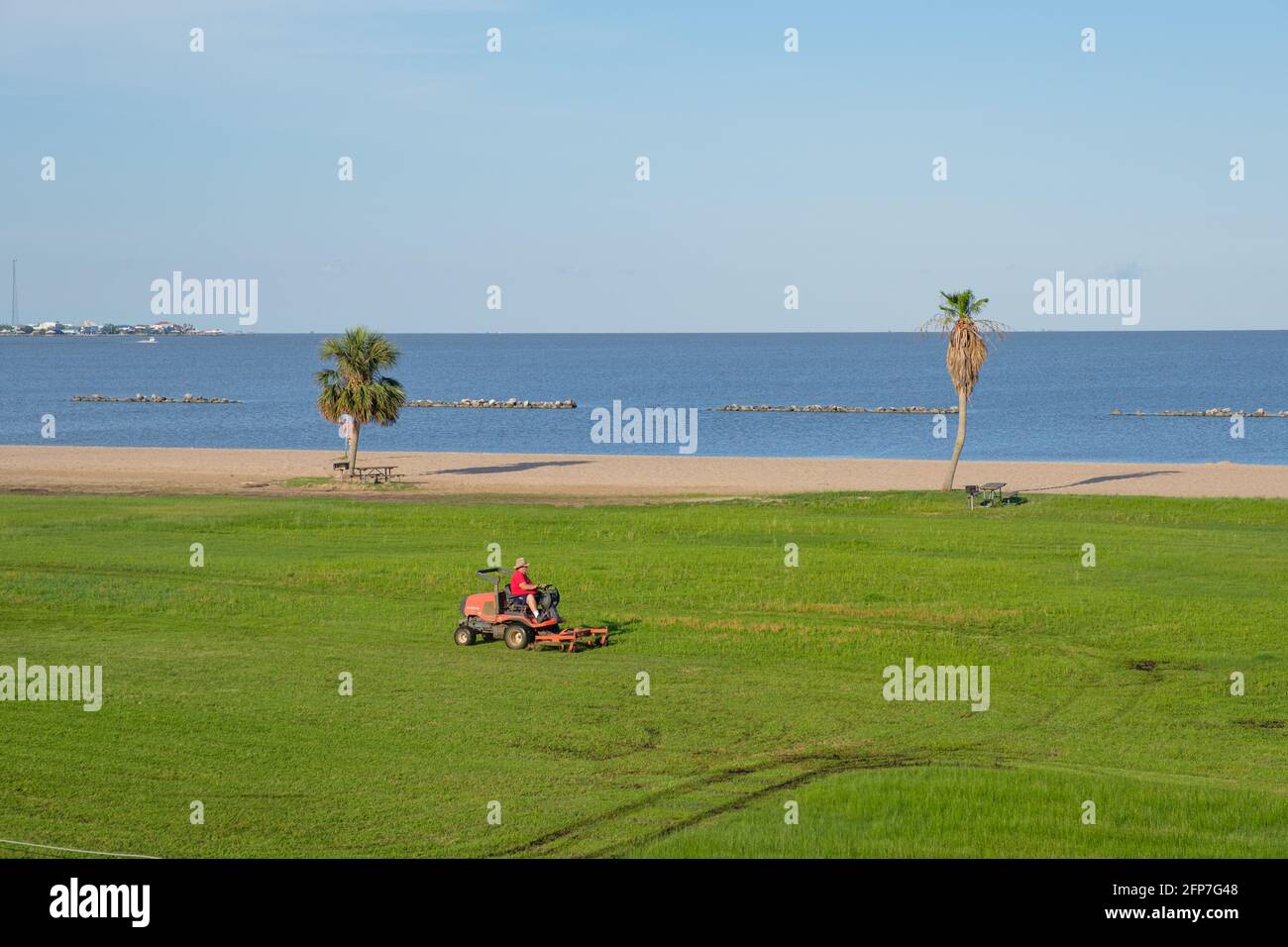 Maintenance worker on riding lawnmower cuts the grass with Vermilion Bay in the background at Cypremort State Park, Louisiana, USA Stock Photo