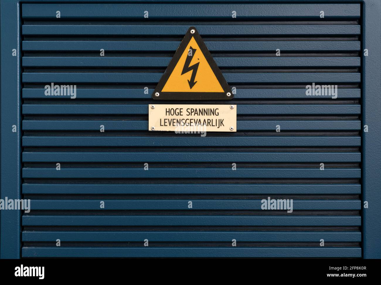 Electrical hazard sign on a triangular yellow sign mounted on a dark blue metal grille with Dutch text 'High Voltage. Dangerous' below it Stock Photo