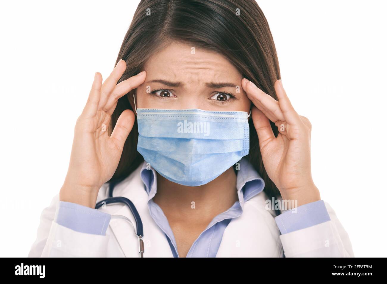 Doctor wearing coronavirus surgical face mask in panic scared doing a funny stressed out facial expression holding head from headache. Asian woman Stock Photo