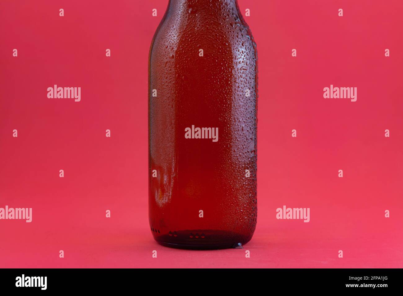 Part of a brown bottle of fresh beer with condensation drops on a red background. The bottle is seen from the middle of the neck down. Stock Photo