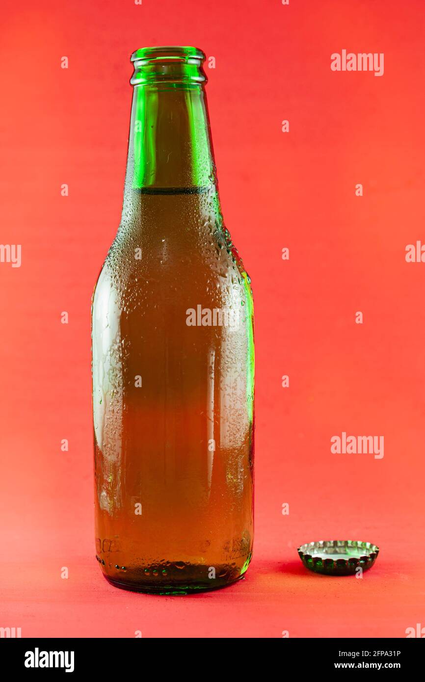 Green glass bottle containing fresh beer with drops of condensation, there is also the cap. The background is red. Stock Photo