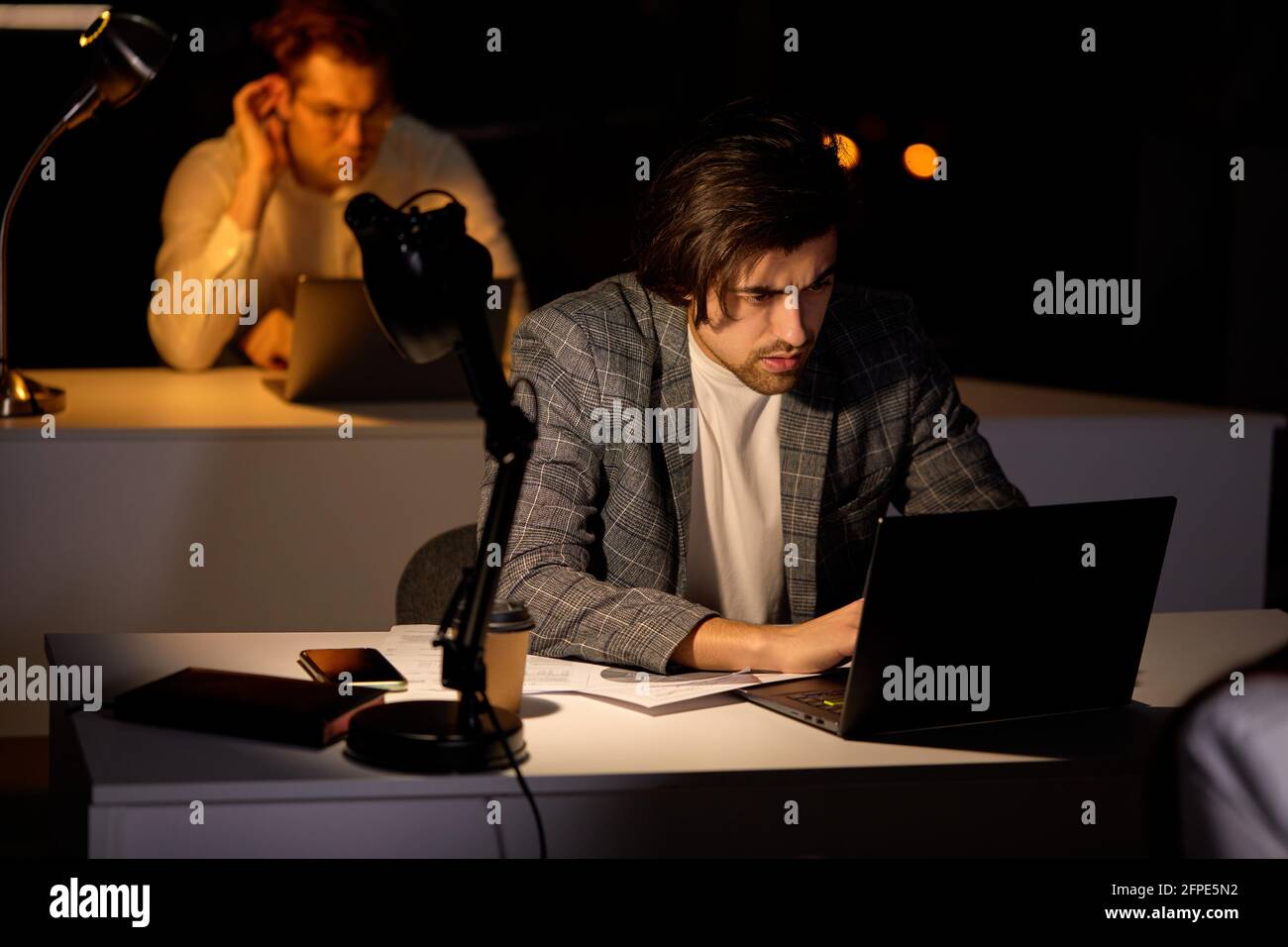 tired focused young man working remotely late at night in dark office using computer, typing, thinking about deadline, in formal wear. Overwork, negat Stock Photo